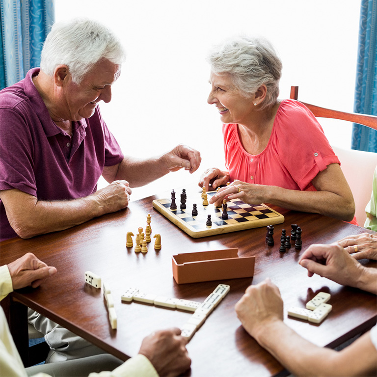 Groupe de personnes âgées souriantes jouant ensemble à des jeux de société, dont les échecs et les dominos, profitant d’un moment convivial dans une pièce lumineuse.