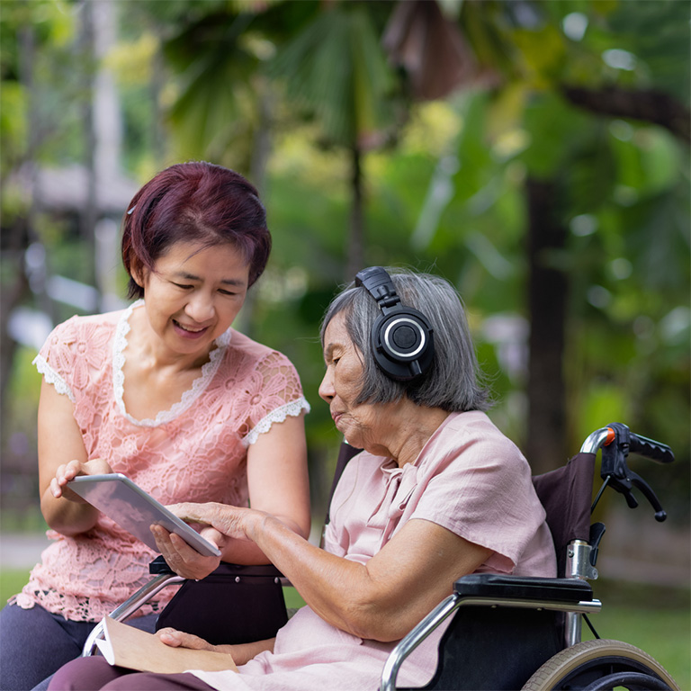Femme âgée en fauteuil roulant portant des écouteurs et écoutant de la musique tandis qu’une autre femme lui montre une tablette, dans un cadre extérieur verdoyant.