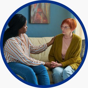 Un préposé aux services de soutien personnel est assis sur un canapé à côté d’une femme âgée, lui tenant doucement la main et posant une main sur son épaule, offrant réconfort et soutien.